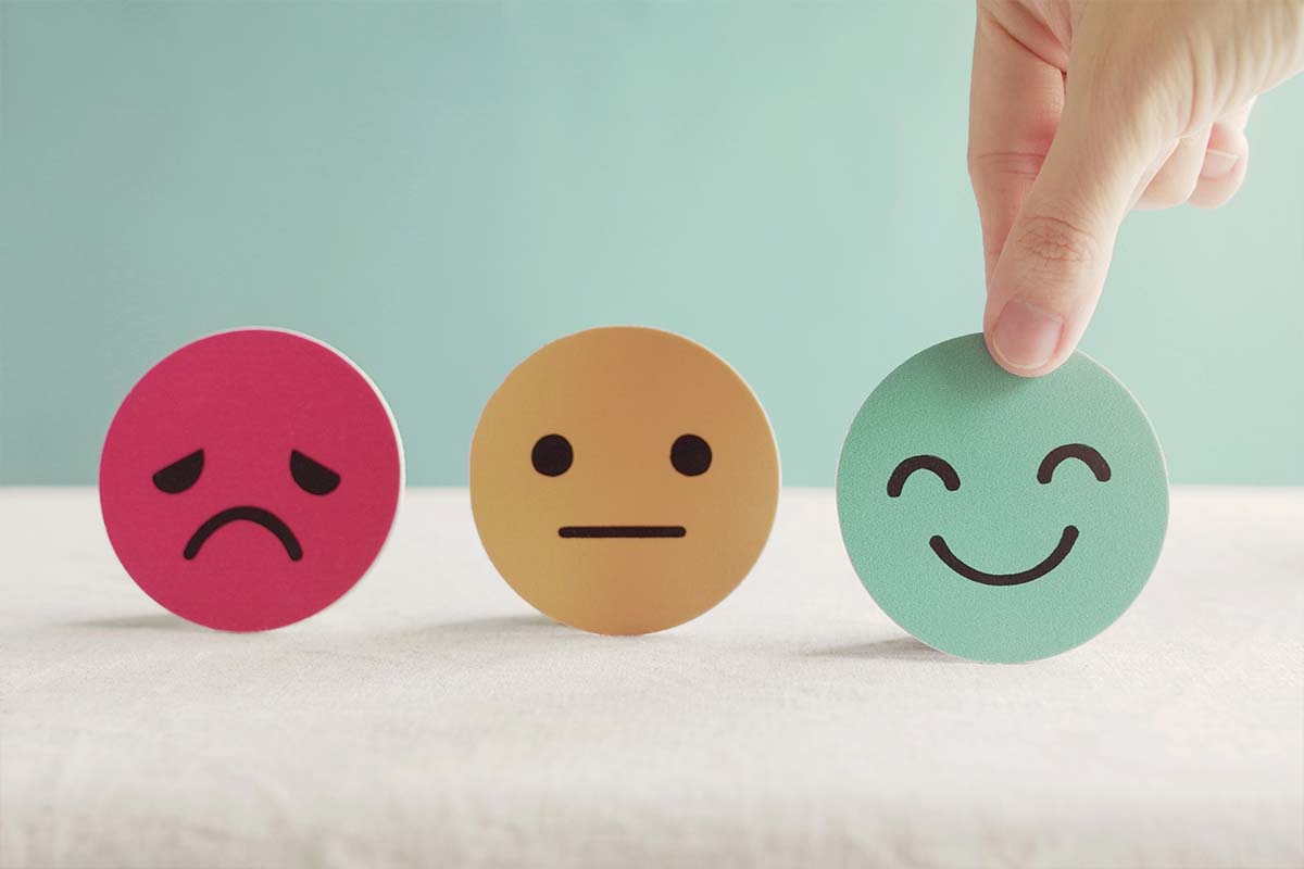 three faces, one sad, one neutral and one happy. Person picking up the smiley face.